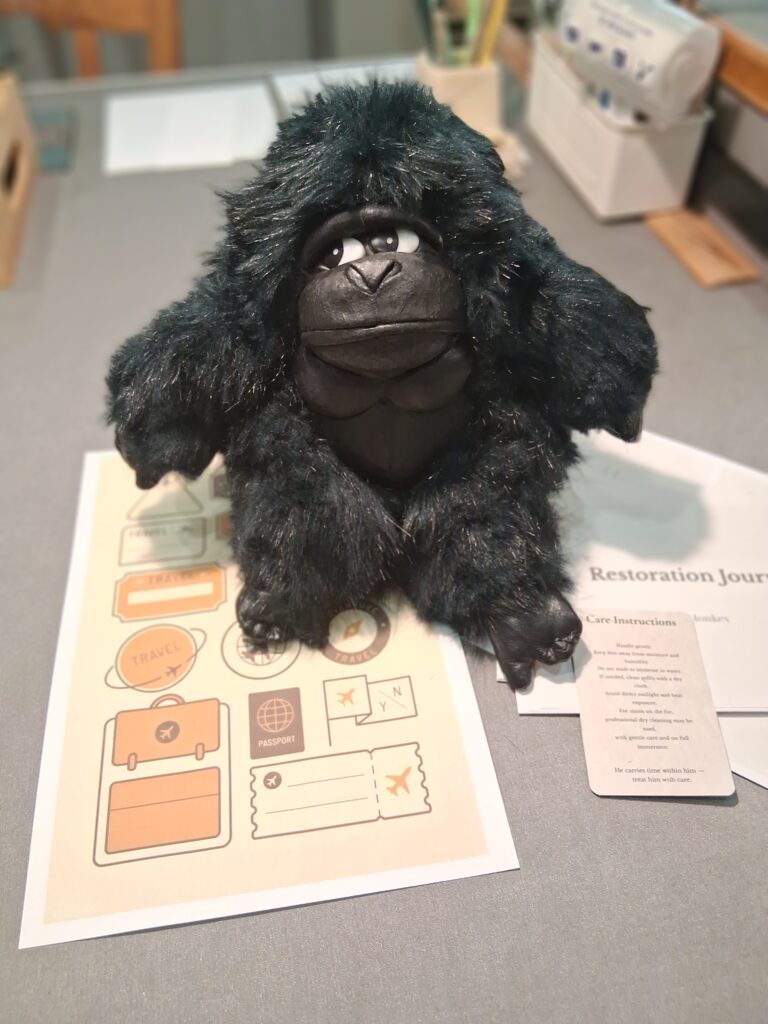 Monkey — Μια Ιστορία Επιστροφής Ο πλήρως αναπαλαιωμένος λούτρινος γορίλας Monkey στο στούντιο Eugenia-Lab, καθισμένος δίπλα στο Restoration Journal, τις κάρτες φροντίδας και τα ταξιδιωτικά έγγραφα.