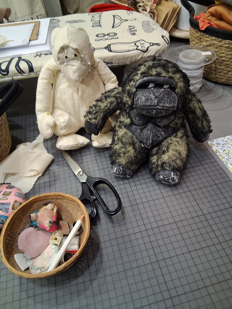 Monkey — Μια Ιστορία Επιστροφής Το λευκό υφασμάτινο ομοίωμα-μακέτα δίπλα στον αυθεντικό λούτρινο γορίλα Monkey, με ψαλίδι και εργαλεία ραπτικής στον πάγκο του εργαστηρίου.